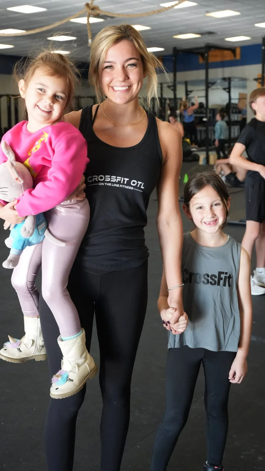 A CrossFit OTL coach holding a toddler and a young girl's hand, all smiling